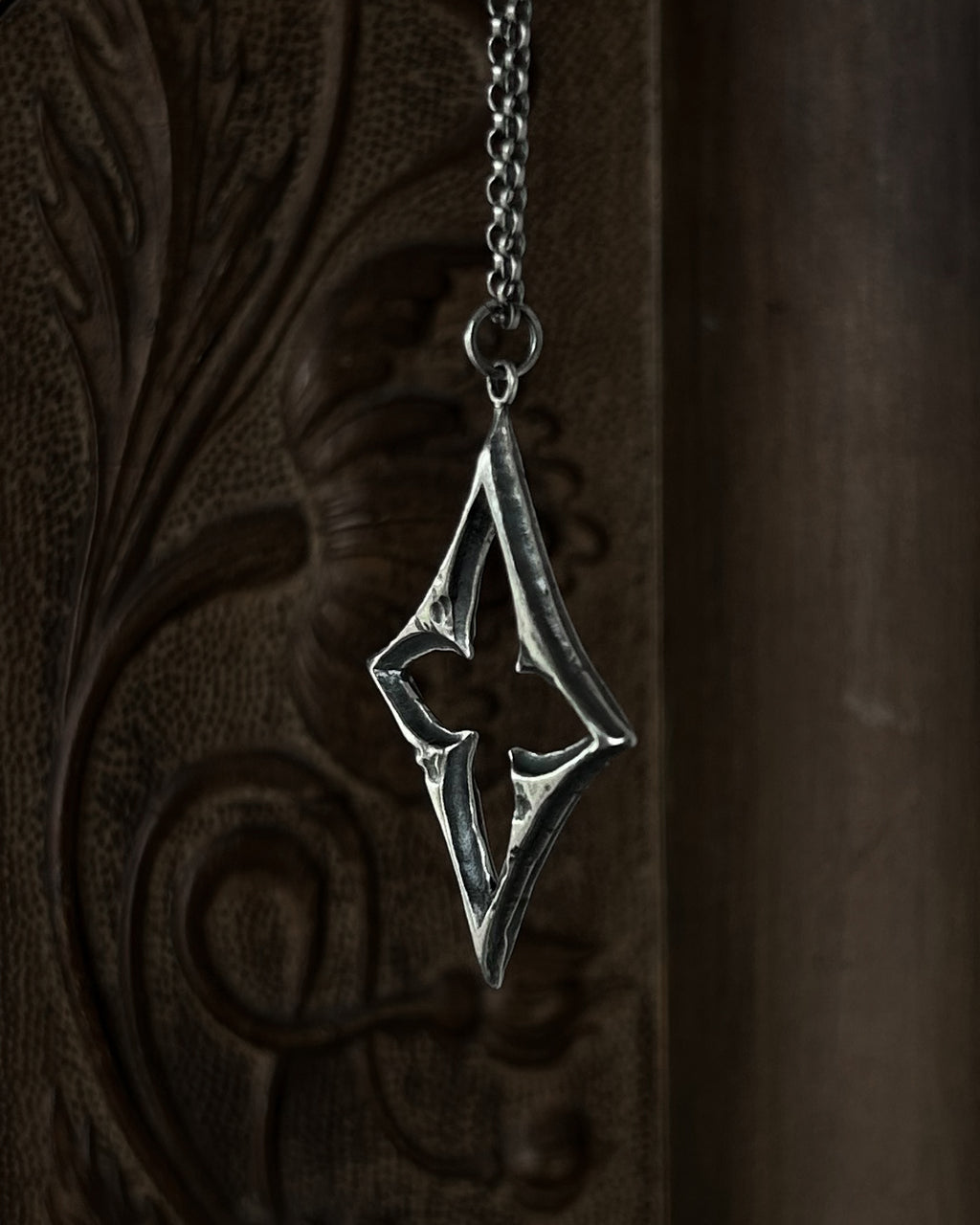 A sterling silver pendant with a stylized open quatrefoil, hanging from a chain against a dark brown carved wooden background.
