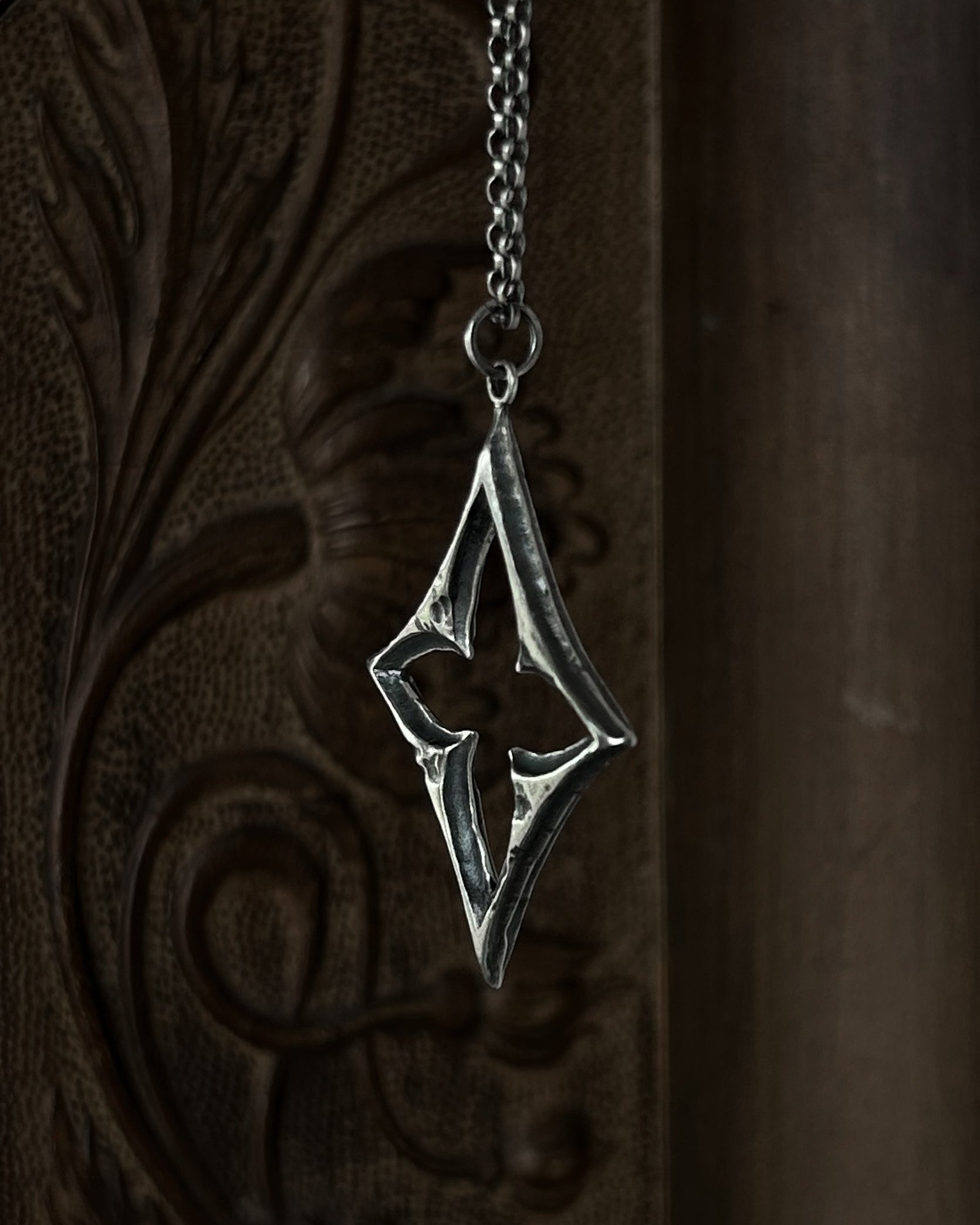 A sterling silver pendant with a stylized open quatrefoil, hanging from a chain against a dark brown carved wooden background.