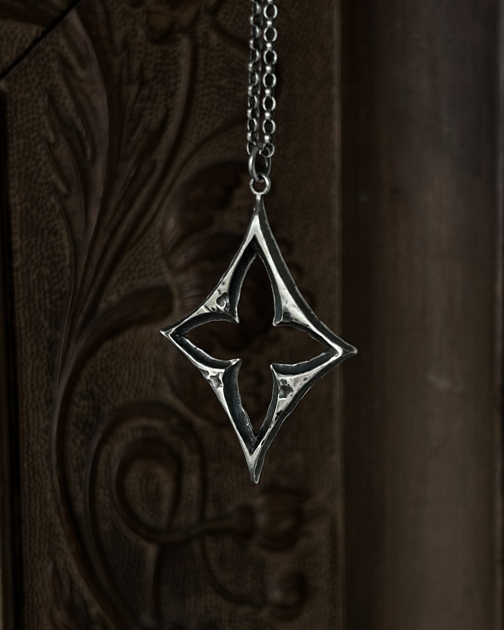 A sterling silver pendant with a stylized open quatrefoil, hanging from a chain against a dark brown carved wooden background.
