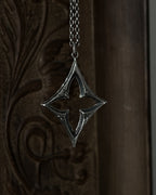 A sterling silver pendant with a stylized open quatrefoil, hanging from a chain against a dark brown carved wooden background.