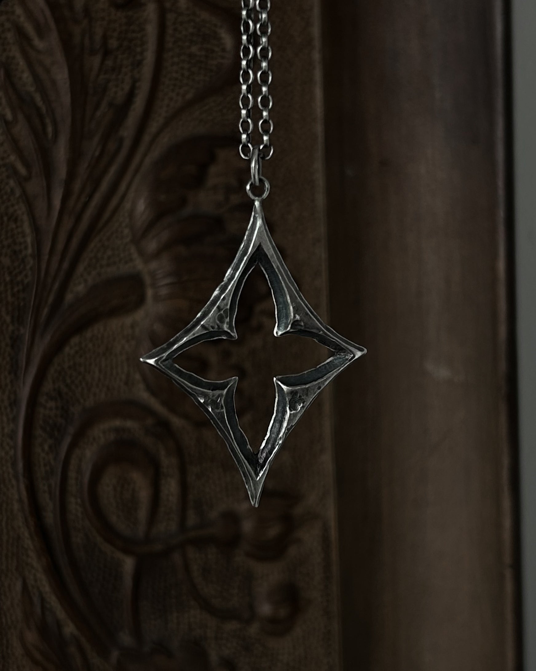 A sterling silver pendant with a stylized open quatrefoil, hanging from a chain against a dark brown carved wooden background.