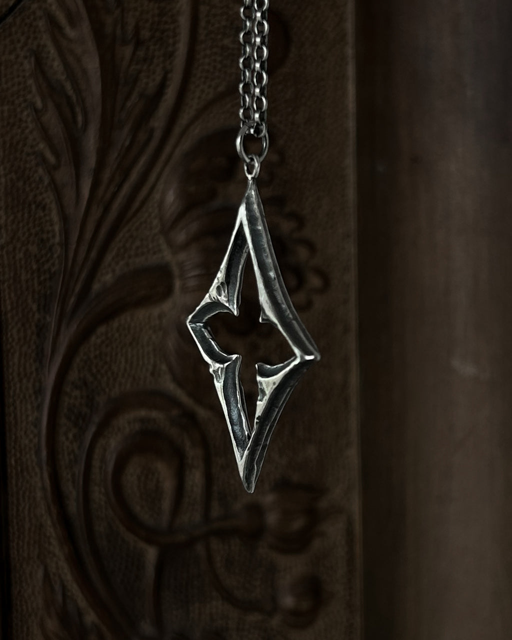 A sterling silver pendant with a stylized open quatrefoil, hanging from a chain against a dark brown carved wooden background.
