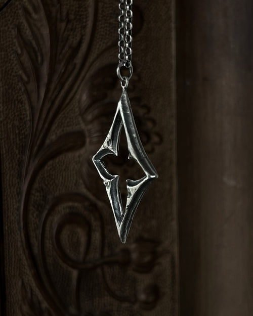 A sterling silver pendant with a stylized open quatrefoil, hanging from a chain against a dark brown carved wooden background.
