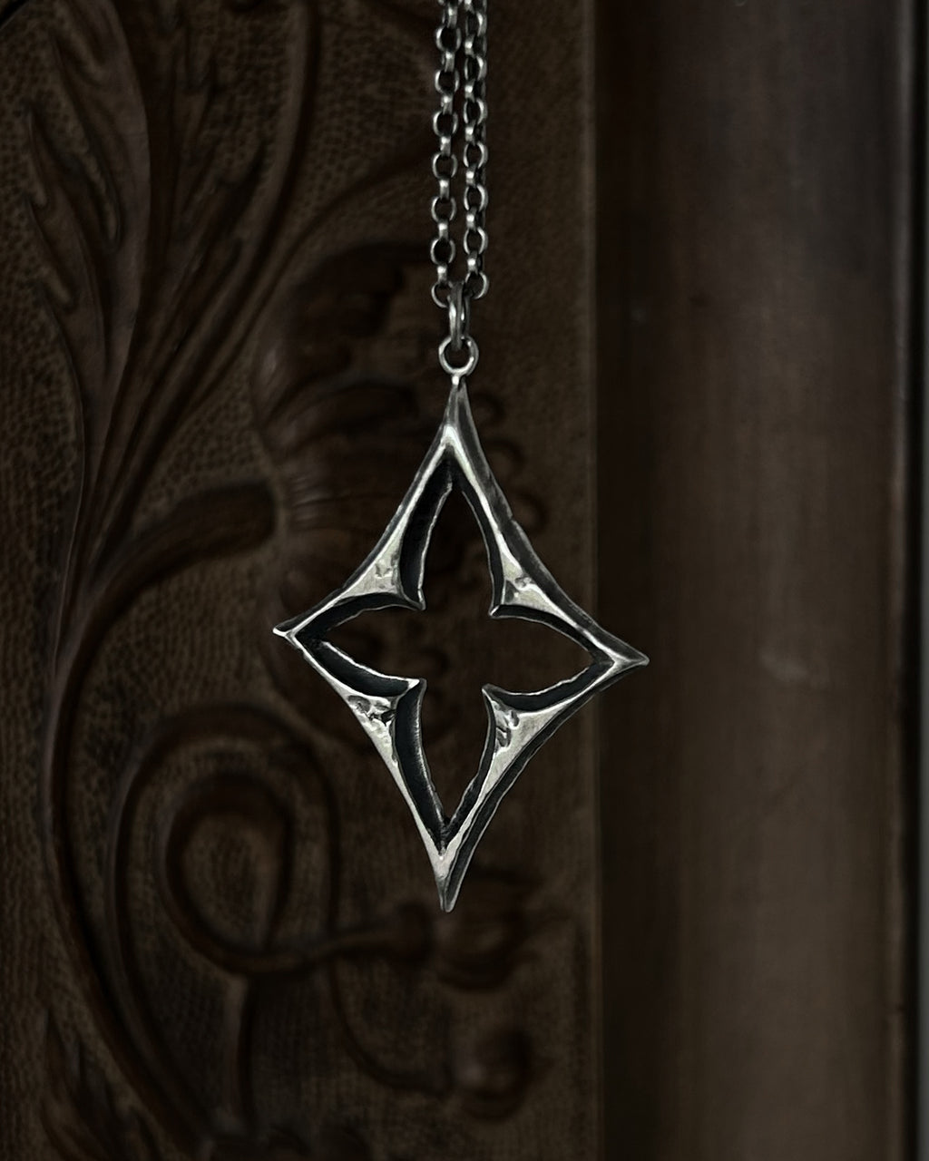 A sterling silver pendant with a stylized open quatrefoil, hanging from a chain against a dark brown carved wooden background.