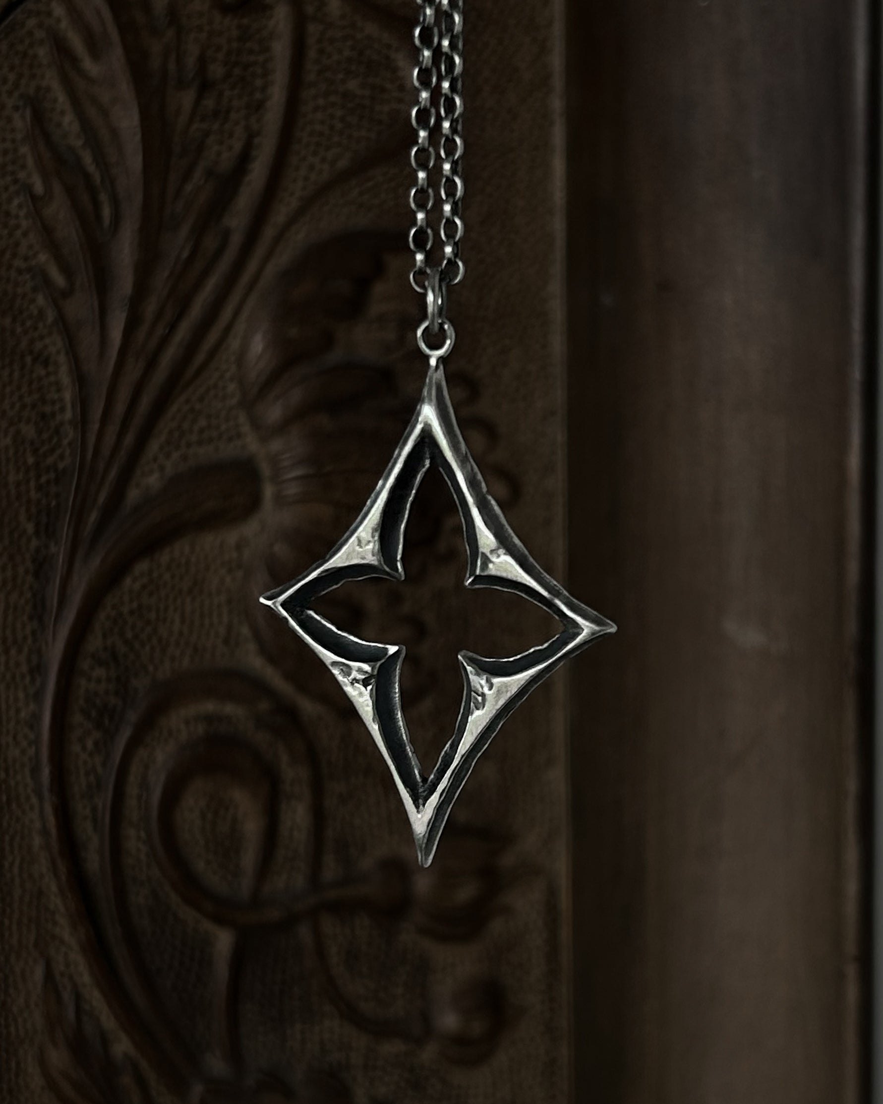 A sterling silver pendant with a stylized open quatrefoil, hanging from a chain against a dark brown carved wooden background.