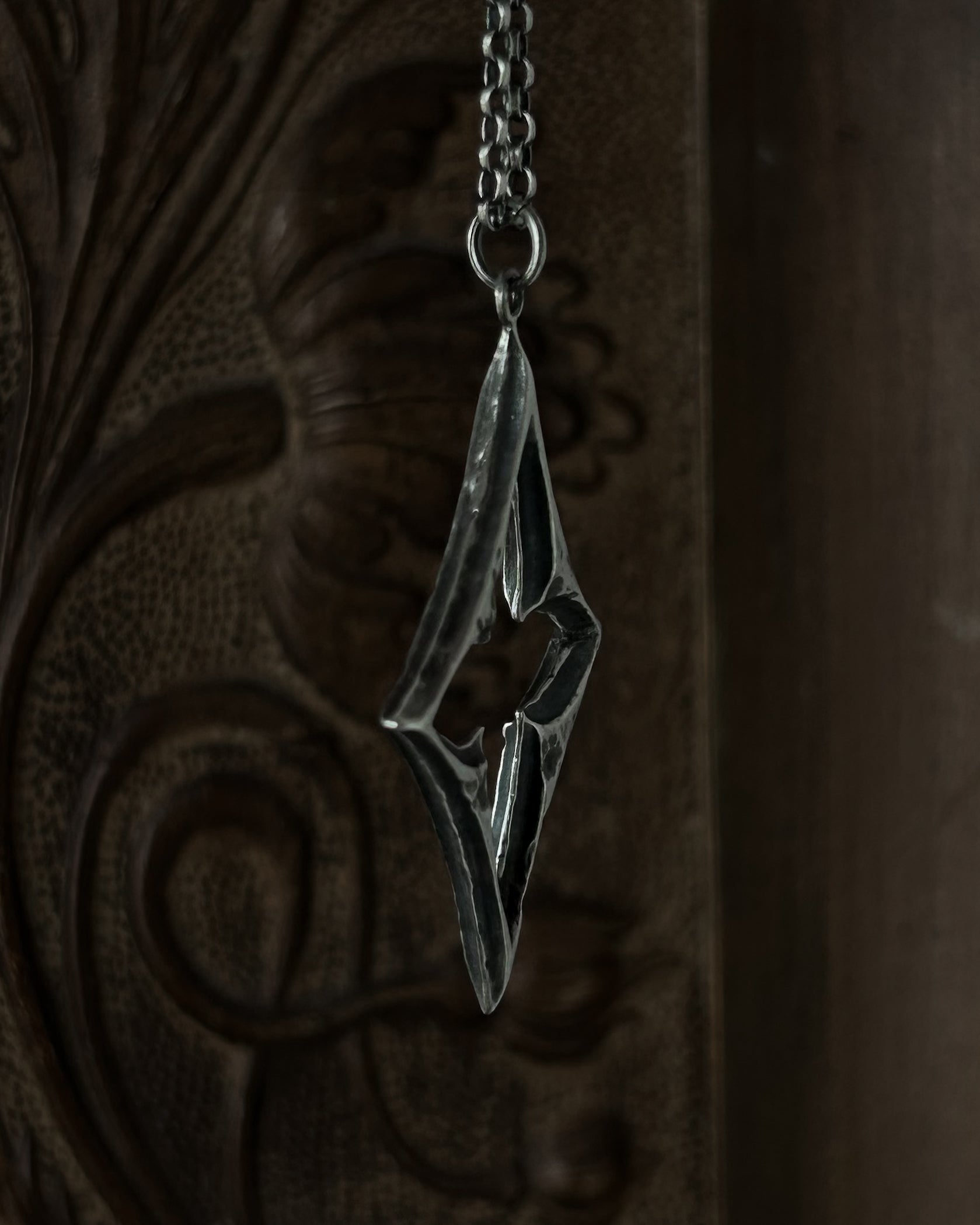 A sterling silver pendant with a stylized open quatrefoil, hanging from a chain against a dark brown carved wooden background.
