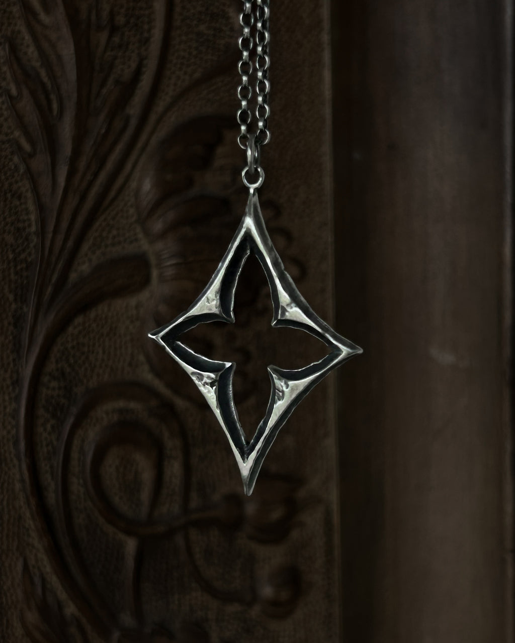 A sterling silver pendant with a stylized open quatrefoil, hanging from a chain against a dark brown carved wooden background.