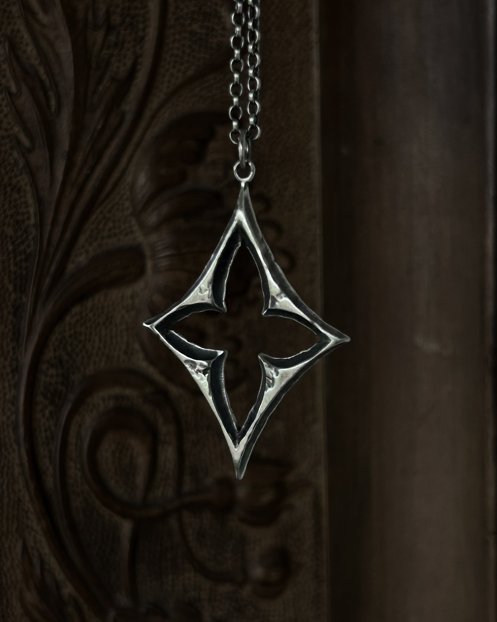 A sterling silver pendant with a stylized open quatrefoil, hanging from a chain against a dark brown carved wooden background.