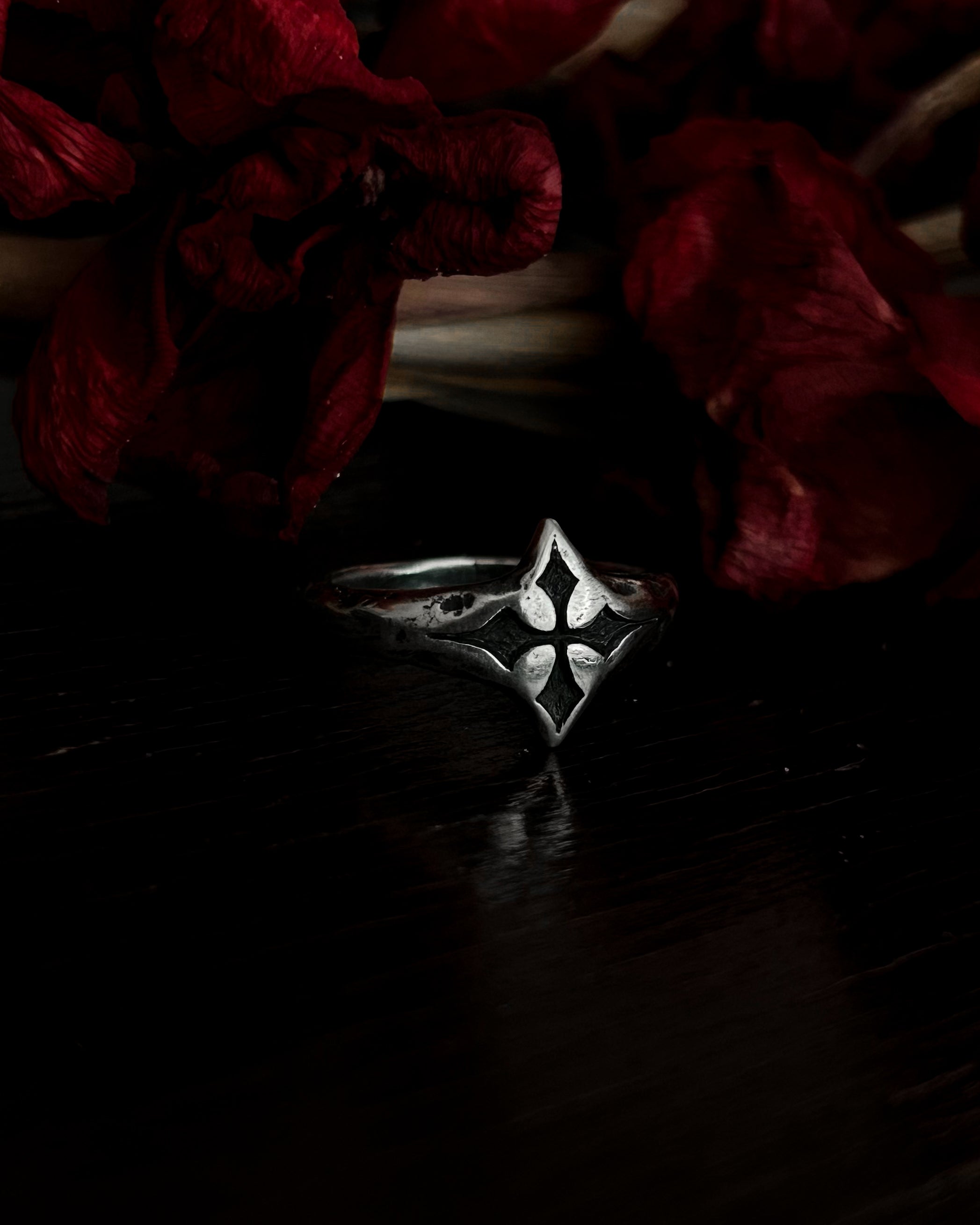 A silver-toned gothic ring with a stylized cross, resting on dark red dried flowers.