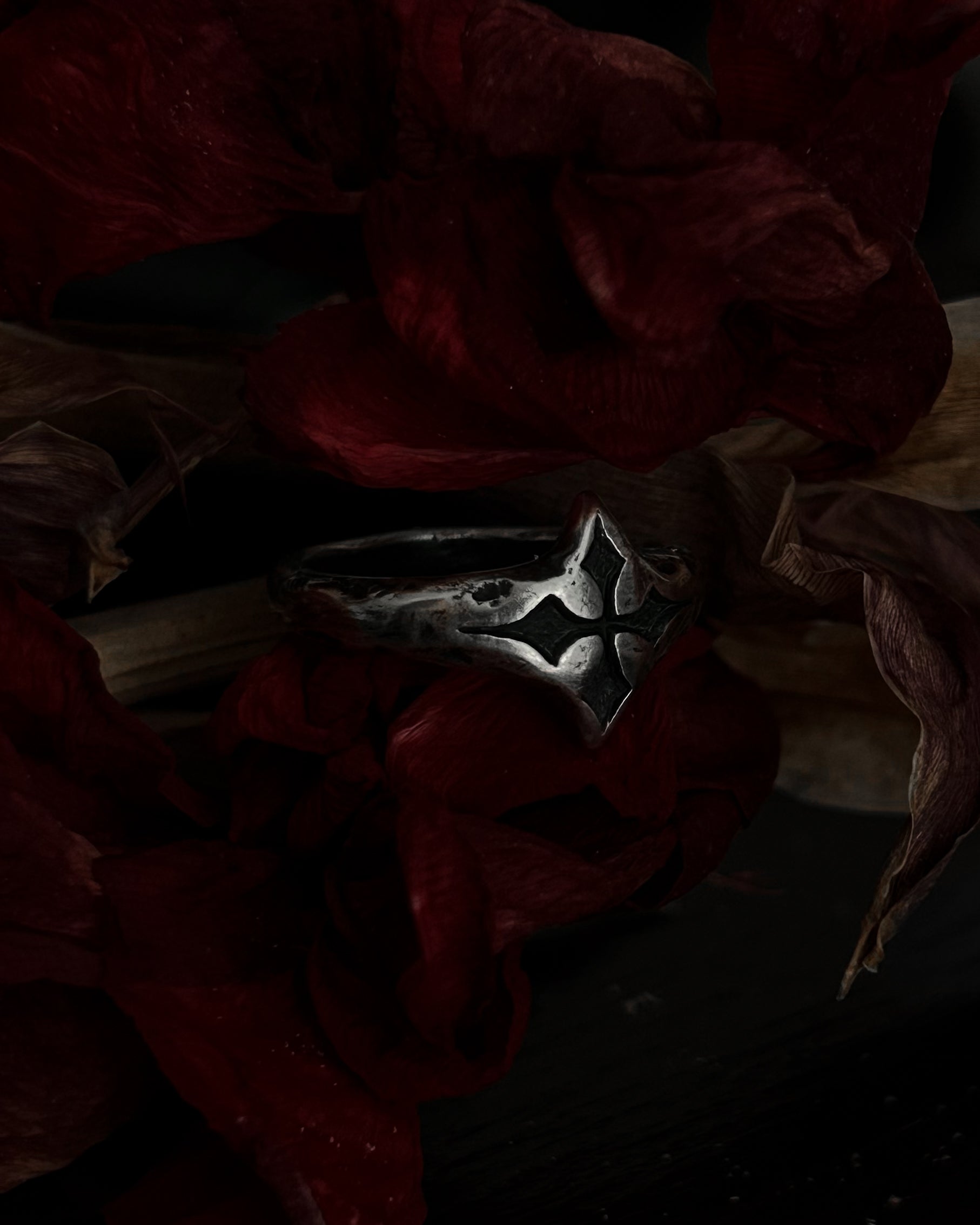 A silver-toned gothic ring with a stylized cross, resting on dark red dried flowers.