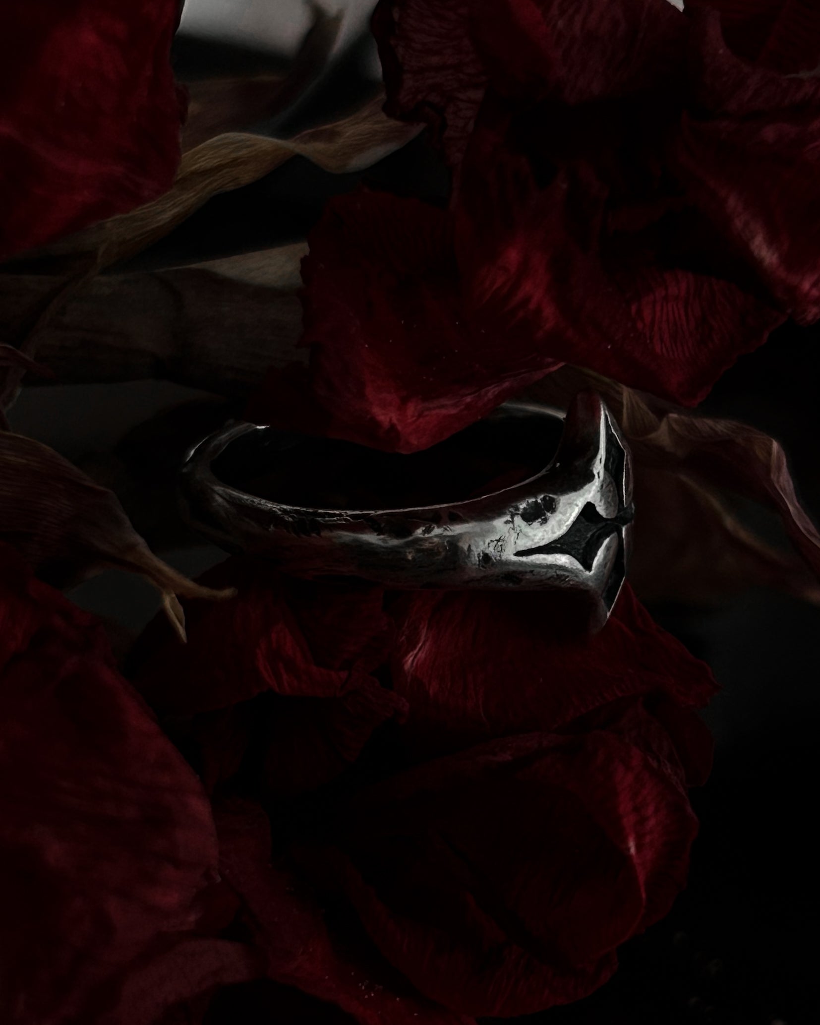 A silver-toned gothic ring with a stylized cross, resting on dark red dried flowers.