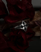 A silver-toned gothic ring with a stylized cross, resting on dark red dried flowers.