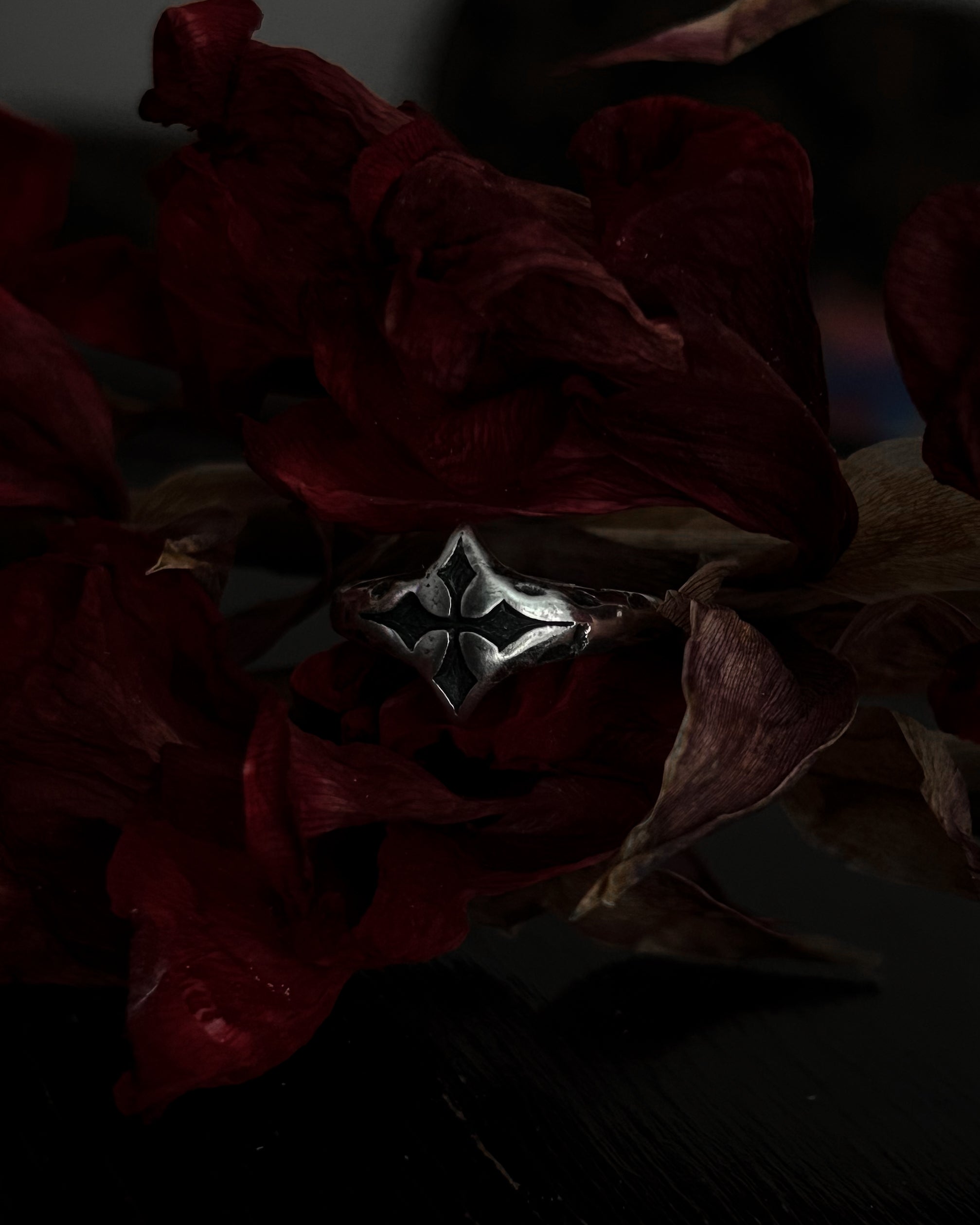 A silver-toned gothic ring with a stylized cross, resting on dark red dried flowers.