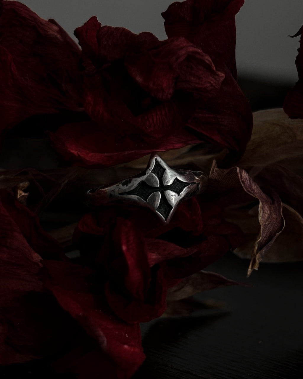 A silver-toned gothic ring with a stylized cross, resting on dark red dried flowers.
