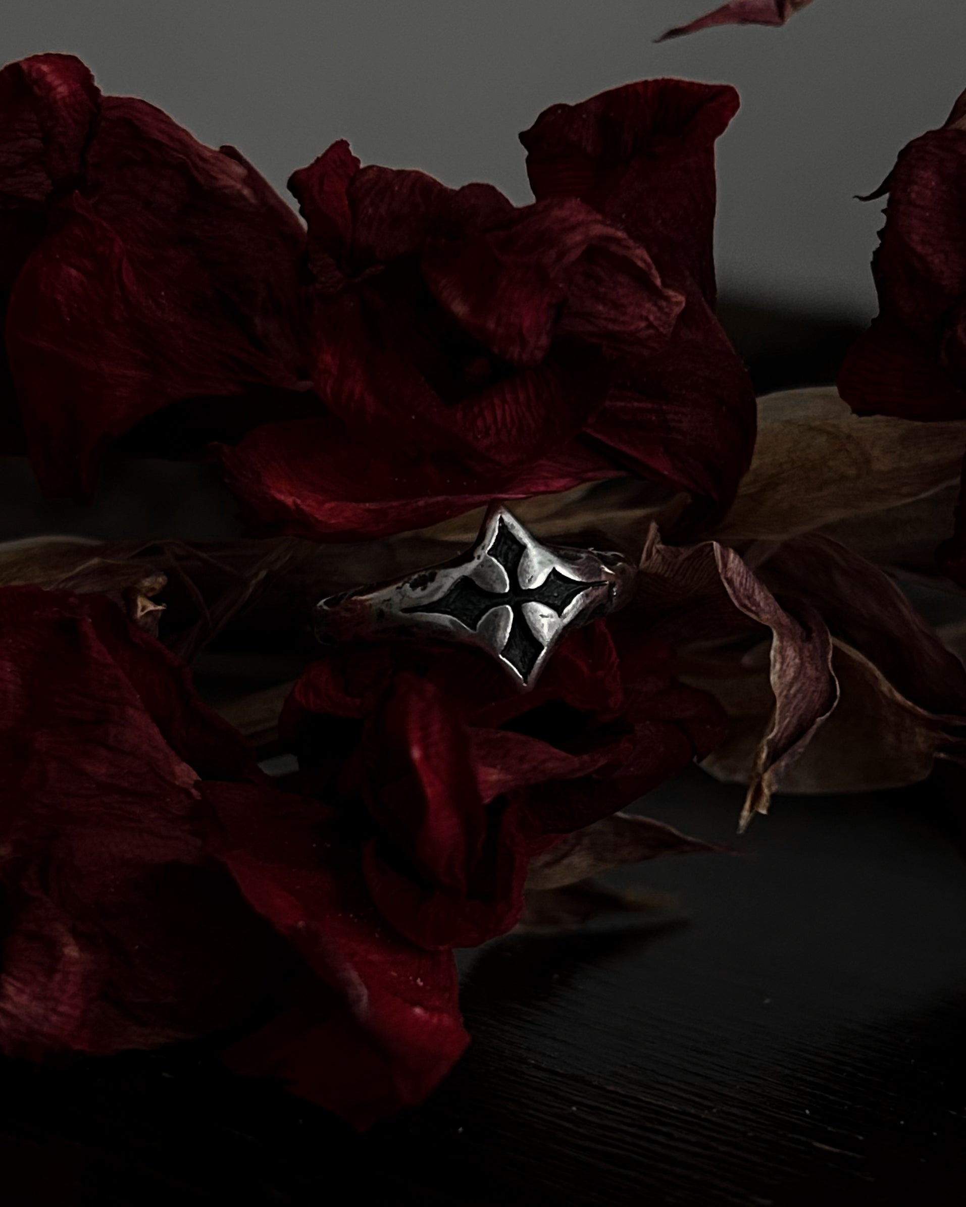 A silver-toned gothic ring with a stylized cross, resting on dark red dried flowers.